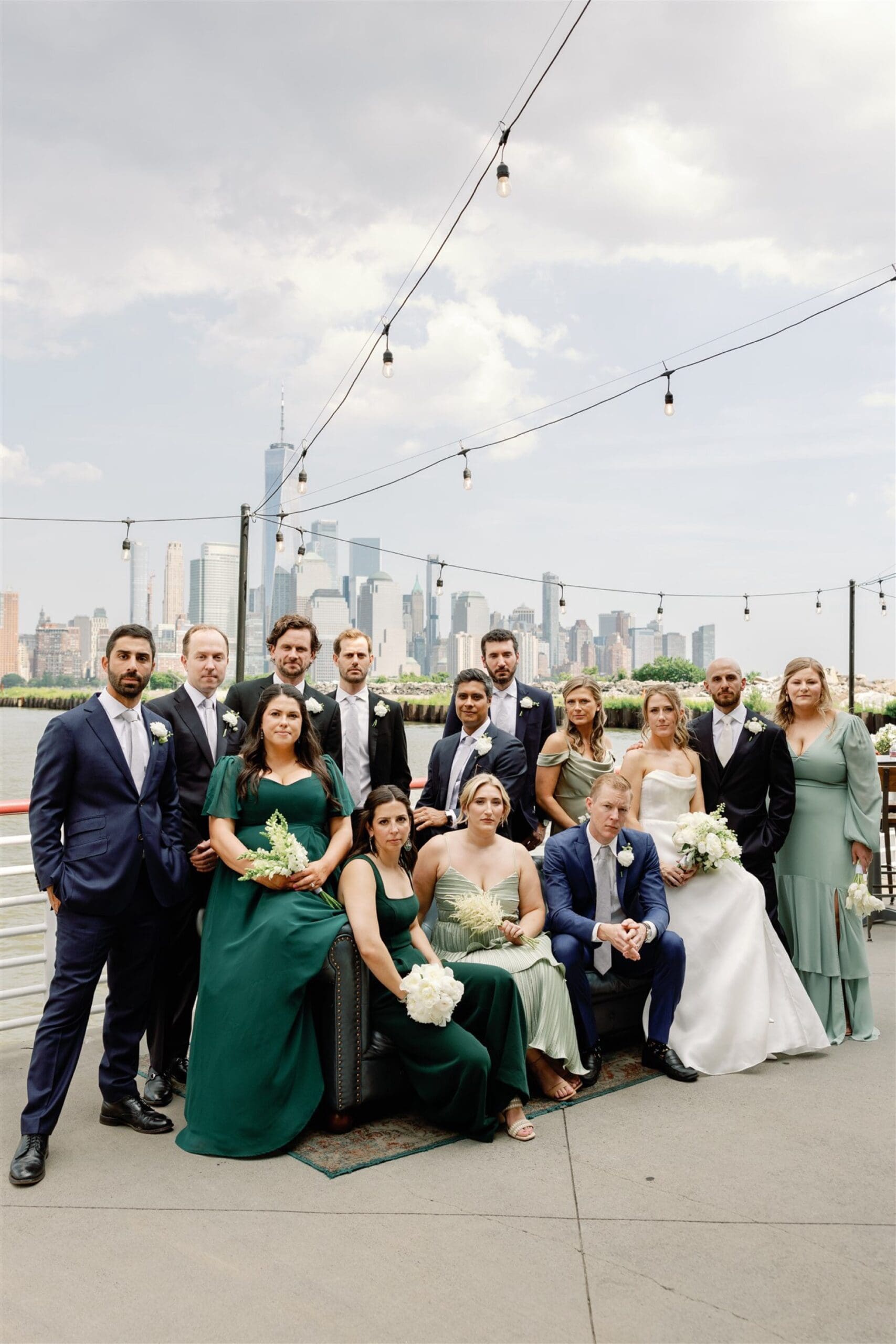 The bride and groom pose with bridal party on a waterfront deck at Battello, with the Manhattan skyline and One World Trade Center in the background.